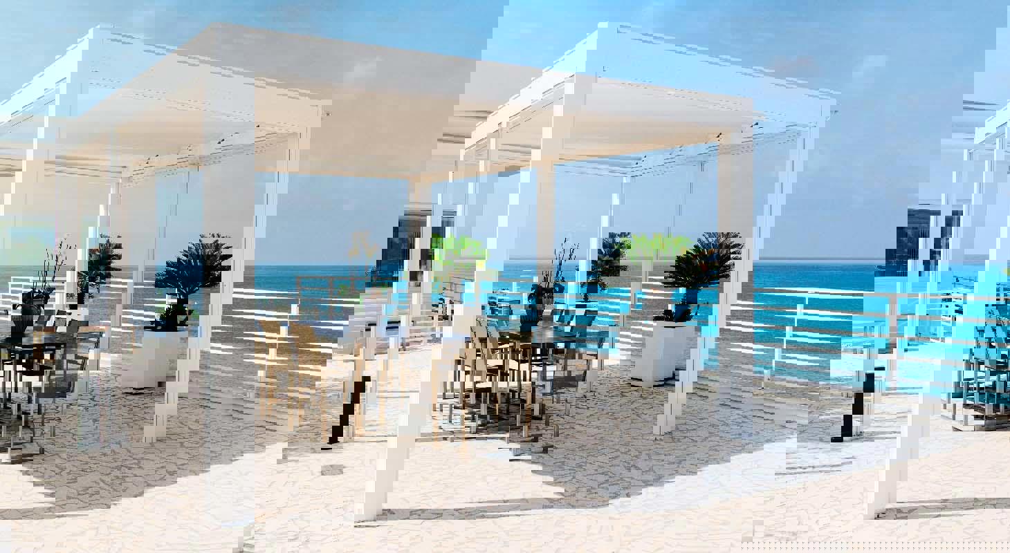 Bela pergola z jedilno mizo in stoli na terasi ob morju, s čudovitim razgledom na ocean in modrim nebom v ozadju.