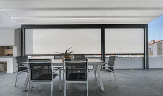 Moderna terasa s pergolo in vgrajenimi screen roloji za zaščito pred soncem, opremljena z jedilno garnituro v črno-beli barvni kombinaciji.