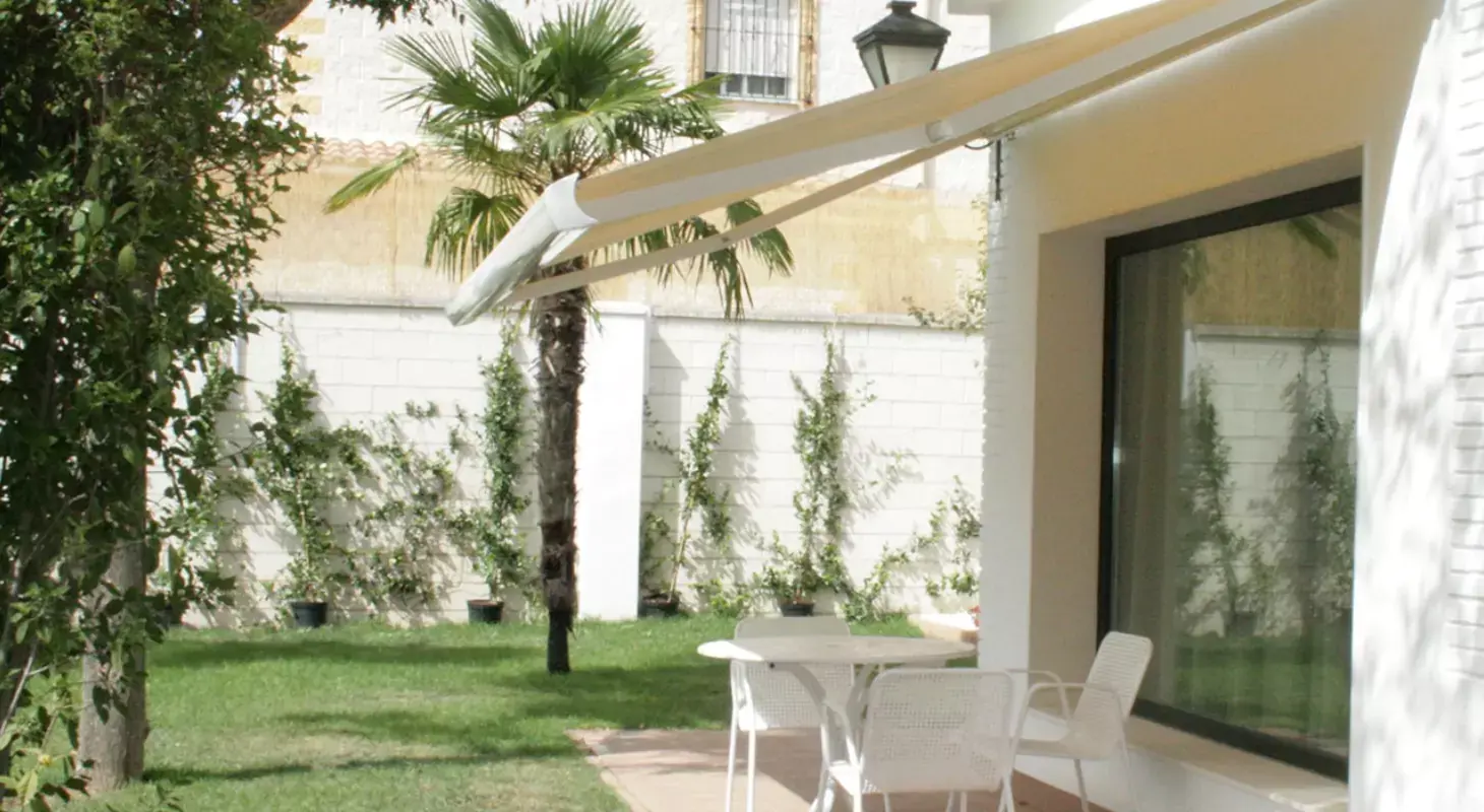 Bež kasetna tenda, nameščena na fasado hiše, ki nudi senco vrtni mizi in stolom na terasi z zelenico in palmami v ozadju.