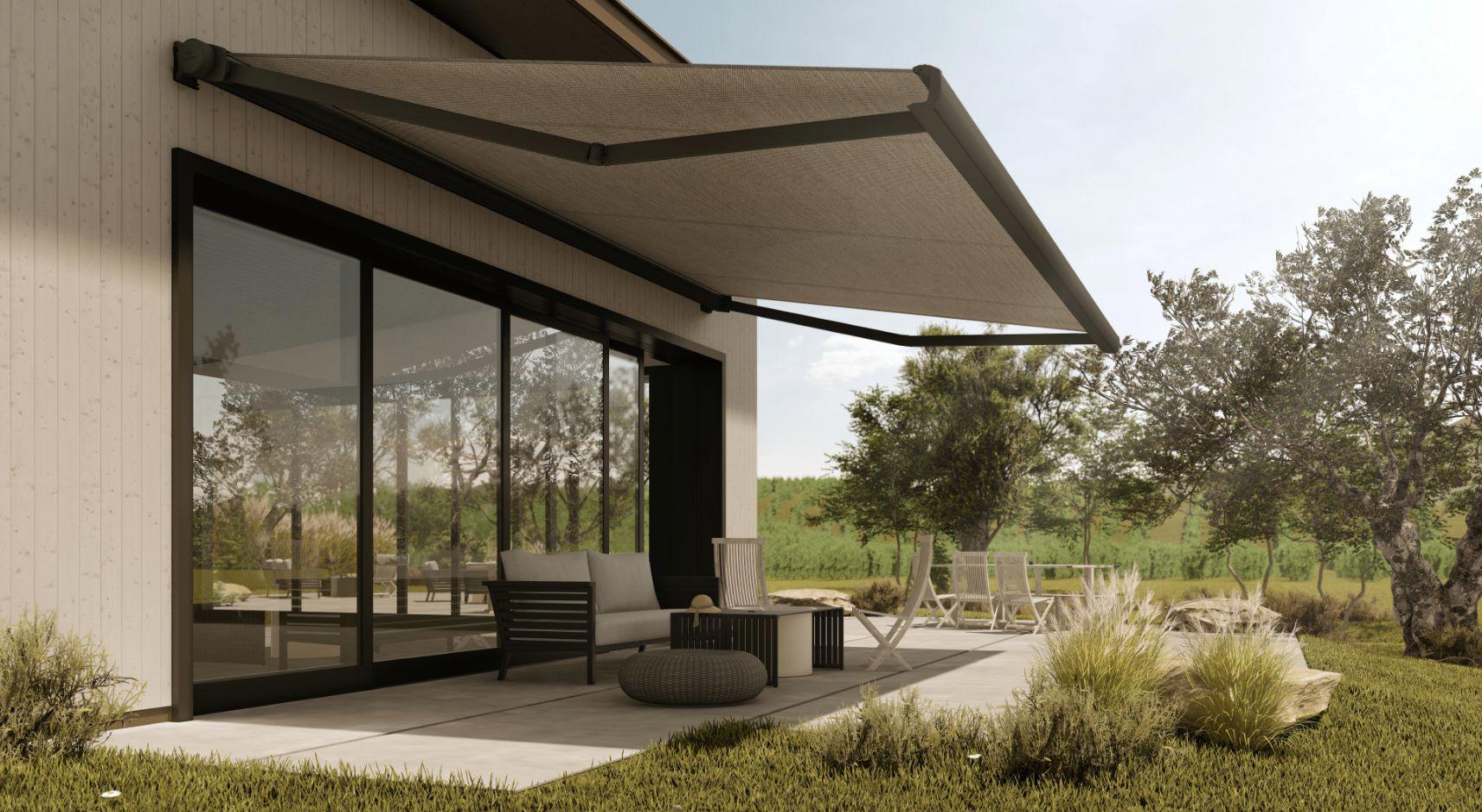 Moderna hiša z veliko stekleno steno in iztegnjeno bež tendo, ki senči teraso z vrtno garnituro, obdano z naravnim zelenjem in drevesi.