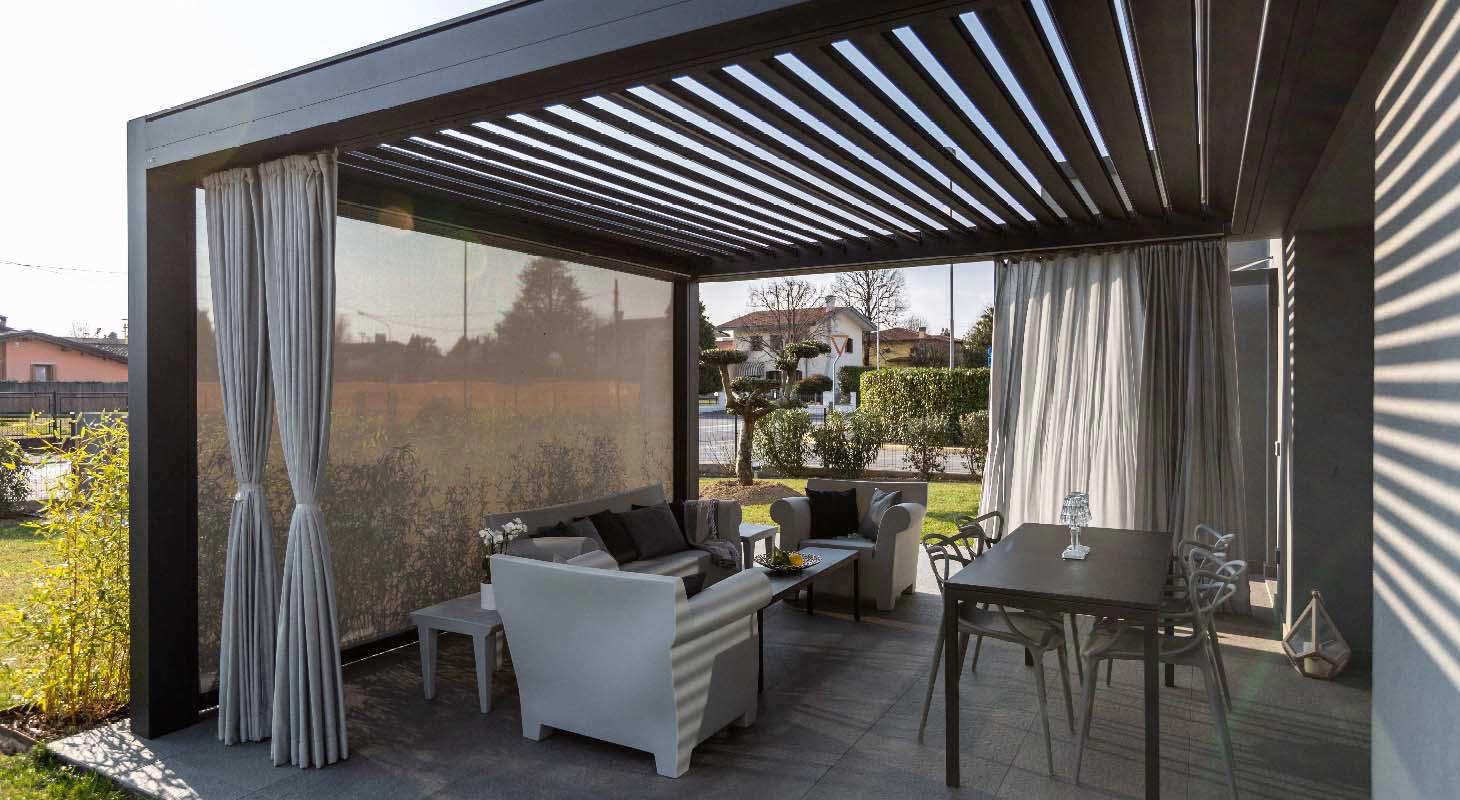 Moderna bioklimatska pergola z nastavljivimi lamelami, zunanjo sedežno garnituro, jedilno mizo in stranskimi zavesami na vrtu ob hiši.