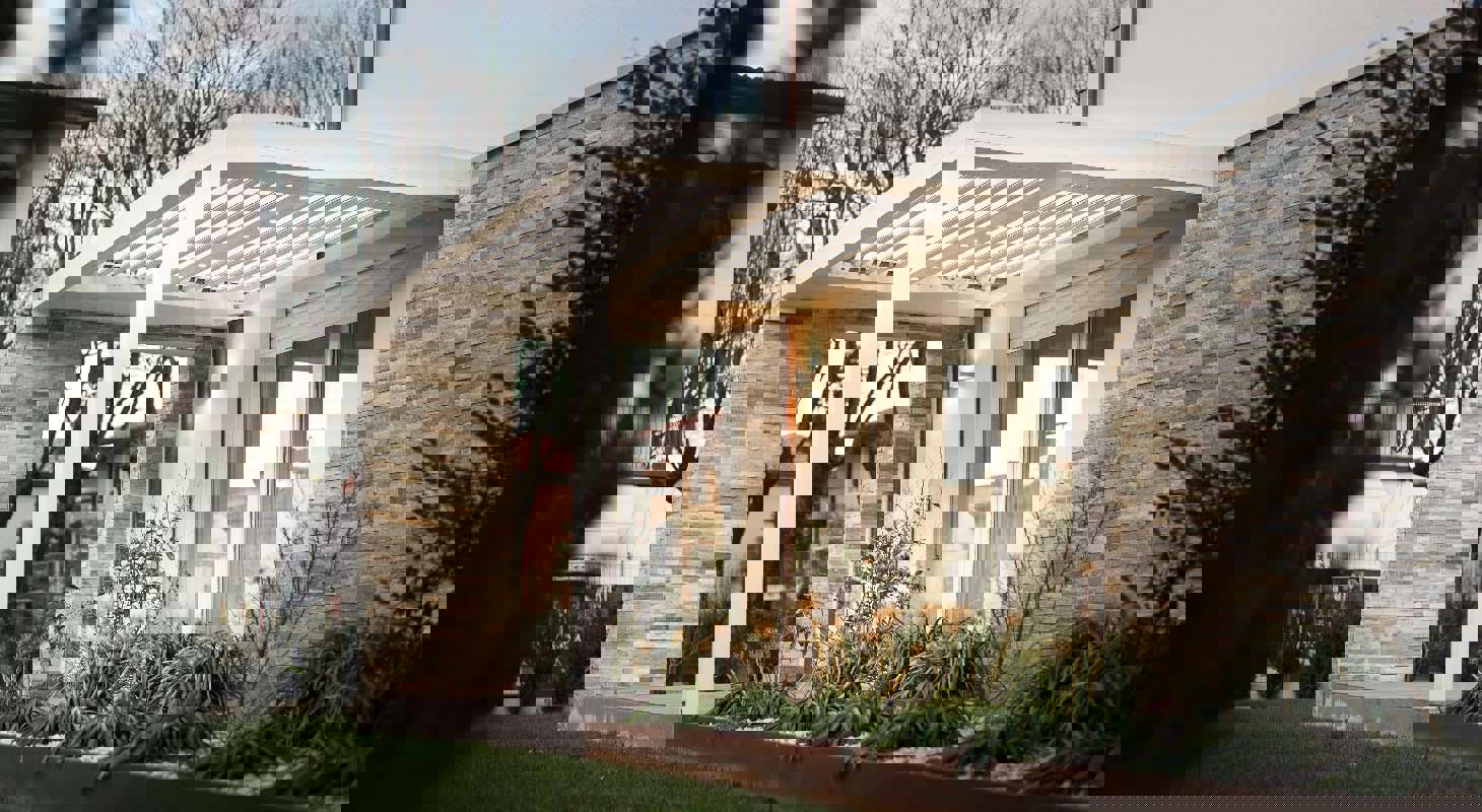 Moderna bela bioklimatska pergola ob družinski hiši z oblogo iz kamna, z integrirano osvetlitvijo in urejeno vrtno okolico.